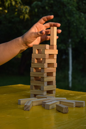 Adult woman builds tower with wooden blocks on yellow table outdoors at sunrise. Balance concept. An...