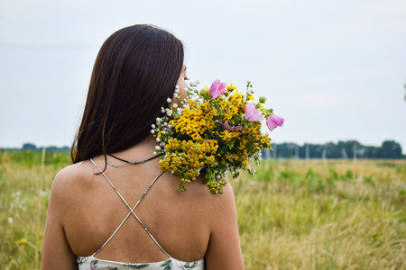 Rear view multiracial young woman holds a bouquet of wildflowers in a picturesque meadow during twilight. The serene scene captures her connection with nature's beauty and the tranquil moments of duskの写真素材