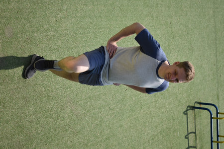 A focused young athlete dynamically performing warm-up drills and stretches outdoors. Clad in...