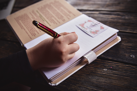 a female hand writing in a notebook with a pen on a wooden tableの写真素材