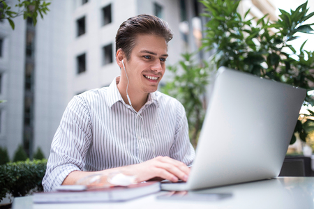 Happy handsome businessman looking at computer screen while typing something on laptop keyboard during work break outdoors, businessman with headphones looking at the notebook sitting in cafe working.の写真素材