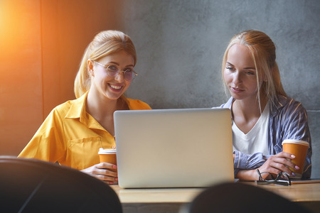 Two cute girls in casual clothes are looking at the screen of a laptop while sitting in office. Bloggers or designers are reading news on a laptop while having coffee break.の写真素材