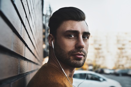 Portrait of smiling man with headphones and cellphone standing by brick wall.の写真素材