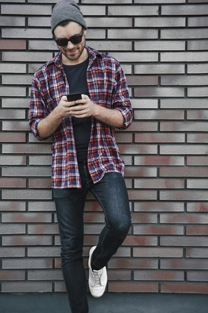 Hipster sms texting phone app in city street on brick wall background. Amazing man holding smartphone in smart casual wear standing. Urban young professional lifestyle.の写真素材