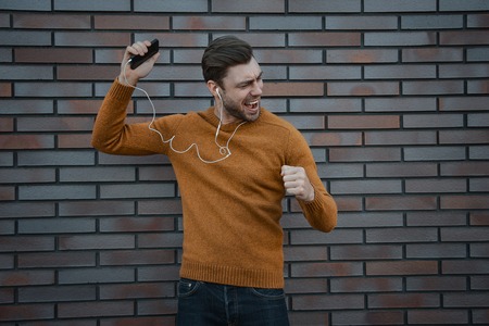 Portrait of smiling man with headphones and cellphone standing by brick wall.の写真素材