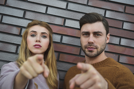 Hey you. Portrait of serious bearded man and woman in casual style standing and pointing at camera with serious face. outdoor shoot, isolated on brick wall background.の写真素材
