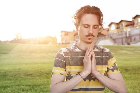 Young man meditating outdoors in the park, sitting with eyes closed and his hands together. man praying to godの写真素材