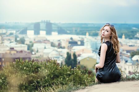 The beautiful lonely girl in black dress and sunglassess sitting on the ground and looking back