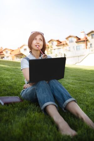Portrait of pretty adult woman sitting on green grass in park with laptop on legs, spending summer day working outdoor, using laptop and wireless Internet for online work. Lifestyleの写真素材