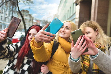 Three beautiful smiling young women walk in the city, using phone outdoors make selfiesの写真素材