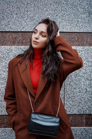 fashionable young woman in a brown coat and red sweater poses against the wallの写真素材