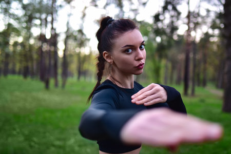 Young woman doing martial arts training in sporty black top holding hands in fighting pose against forest background.の写真素材