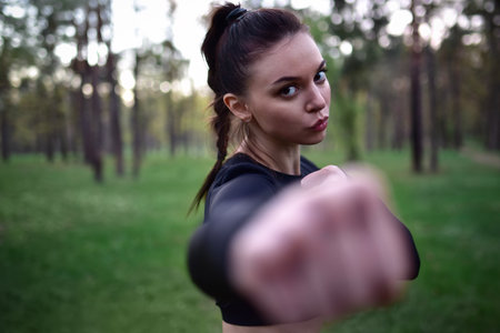 Young woman doing martial arts training in sporty black top holding hands in fighting pose against forest background.の写真素材