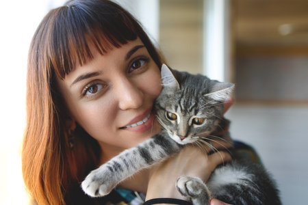 A nurturing woman tenderly cradles a striped gray kitten close to her chest, her warm expression capturing a heartfelt moment of care in a softly blurred indoor setting.の写真素材