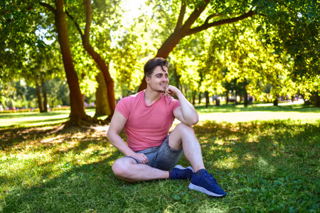 A happy young man sits alone on the grass in a sunny park, looking away and smiling while resting his head on his hand. The image conveys a feeling of contentment and relaxation outdoorsの写真素材