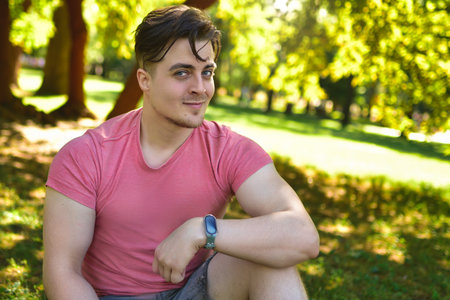 A close-up portrait captures a young man sitting on the grass in a park, looking at the camera with a pleasant, subtle smile. The soft focus background highlights his expression and the sunny outdoor environment.の写真素材