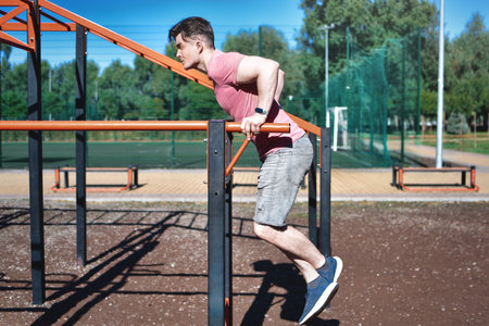A young man is shown performing dips on parallel bars at an outdoor workout area on a sunny day. He is focused on his exercise, demonstrating strength and fitness in a park environment.の写真素材