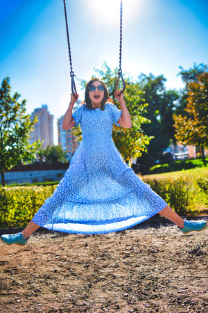A joyful young woman is energetically swinging high on a swing set in a sunny park. She is laughing and spreading her legs wide, embodying a feeling of freedom, happiness, and playful abandonment in the bright outdoorsの写真素材