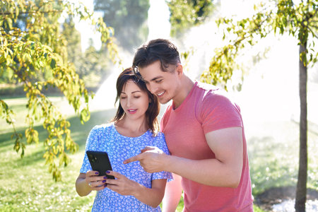 Two happy young friends are standing in a sunny park with active sprinklers, looking together at a smartphone. They appear engaged and joyful, sharing a moment while blending technology use with outdoor leisure in a refreshing environment.の写真素材