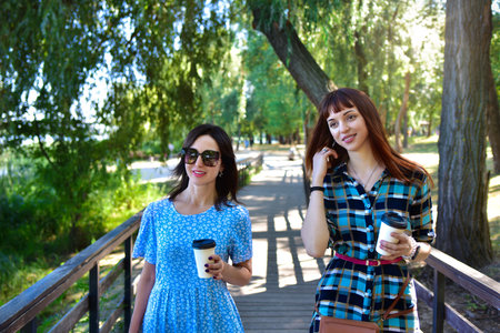 Two cheerful women stroll along a wooden pathway in a sunlit park, each holding a coffee cup and sharing a relaxed moment amid lush greenery. One wears a blue floral dress, while the other sports a blue-green plaid shirt dress with a red belt and carries a brown purse, contributing to the inviting, joyful vibe of the sceneの写真素材