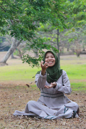 portrait of asian woman playing in the park. Muslim model wearing a robe is sitting on the grass with a happy expressionの写真素材