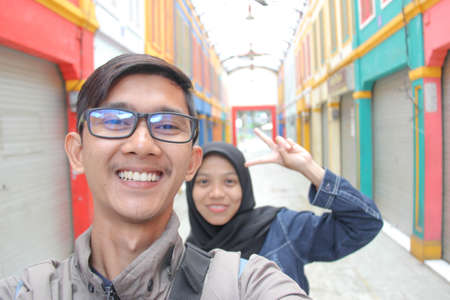 portrait of an Asian man and woman taking a photo together holding a camera against a market background. togetherness in a romantic and happy relationship while on vacationの写真素材