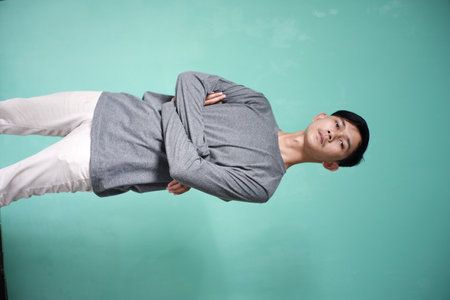 portrait of handsome asian man wearing gray t-shirt with cool pose. skinny man on green background. man folds hands on stomach with serious expressionの写真素材