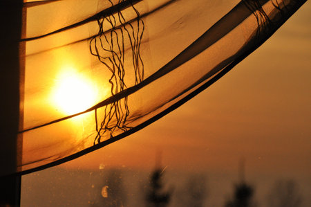 The setting sun shines through the window curtain, casting beautiful warm orange lightの写真素材