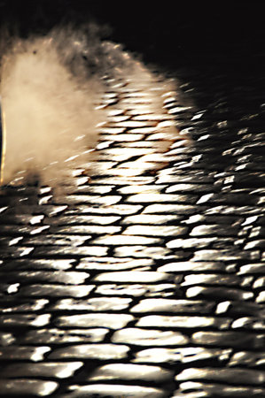 Cars drive on a cobblestone street at night, car lights on the streetの写真素材