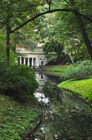 A beautiful view showcases a historic white building with elegant columns, framed by the vibrant colors of autumn trees in Warsaw's Royal Åazienki Park. A gentle stream leads towards the building, adding a touch of tranquility to the scene. The image captures the harmonious blend of architecture and nature, ideal for themes related to travel, history, parks, and autumn beauty.の写真素材