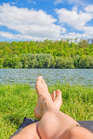 male feet at lake - man relaxing at lake on camping mat - green forest / trees in backgroundの写真素材