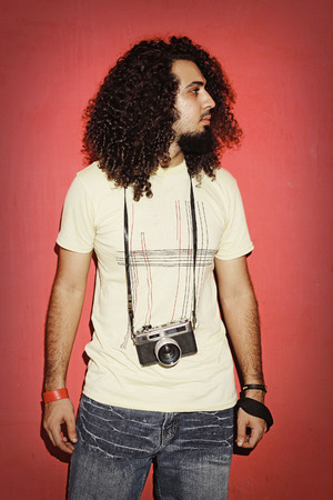 Closeup of one handsome passionate expressive cool young brunette photographer men with long curly hair holding a vintage SLR camera standing against red backgroundの写真素材