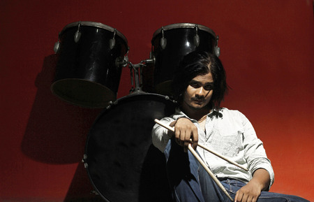 young drummer holding sticks and sitting next to drum kit in spot light on red background looking into the cameraの写真素材