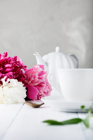 Fresh beautiful flowers peonies and a ceramic white cup with tea for breakfast on an old wooden tableの写真素材