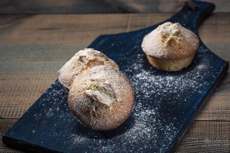 Homemade fresh muffins sprinkled with icing sugar on a wooden board. Dark background.