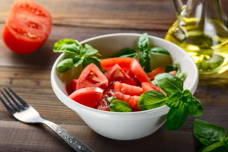 Vegetarian food. Fresh red tomatoes are sliced in a white ceramic plate with basil herb and olive oil on a wooden table.の写真素材
