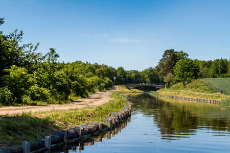 Summer European landscape. Canal among trees, shrubs and fields, dirt road along the canal, bridge over the canal. Elblag Canal, Poland. Horizontalの写真素材