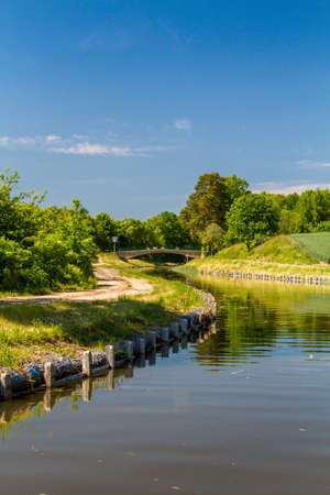 Summer European landscape. Canal among trees, shrubs and fields, dirt road along the canal, bridge over the canal. Elblag Canal, Poland. Verticalの写真素材