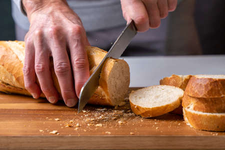 hands cutting a freshly baked baguette into slices on a wooden board, fron view.