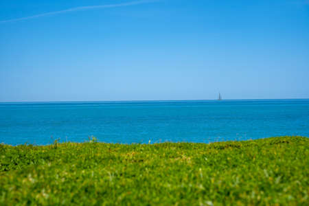 Blue sky seascape with sailboat on the horizon. View from the green hill of an island.の写真素材