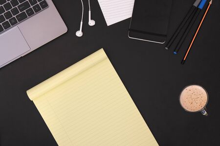 Office work table with coffee, supplies, pencil, pens, smartwatch, earphones, notepad, and laptop. Top view with a legal notebook with a space for your text.の写真素材