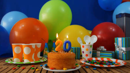 Birthday cake on rustic wooden table with background of colorful balloons, gifts, plastic cups and plastic plate with candies and blue wall in the backgroundの写真素材