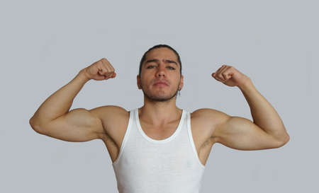 Young hispanic man with hair pulled up bun in white sleeveless t-shirt showing muscles of his arms looking towards cameraの写真素材