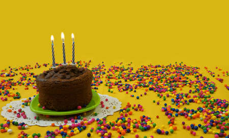 Chocolate birthday cake with 3 blue and white candles lit, on a small green plate, surrounded by candy balls, scattered on a yellow background, Image with copy spaceの写真素材