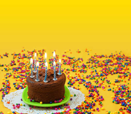 Chocolate birthday cake with lighted blue and white candles, on a small green plate surrounded by candy balls strewn on a yellow background. Image with copy spaceの写真素材