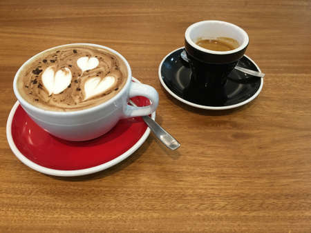 Two ceramic cups with hot coffee drink in red and black dishes on a wooden table with a metal spoon