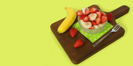 Healthy breakfast Glass plate with chopped strawberries and pieces of banana on a green paper napkin on a dark wooden board, decorated with 3 strawberries, a banana with peel and a fork on green backgroundの写真素材