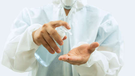 Doctor with mask and white bioprotective suit washing his hands with a bottle of alcohol gel in the foreground on white backgroundの写真素材