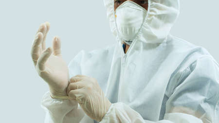Doctor with mask and white bioprotective suit putting latex gloves on hands on white background