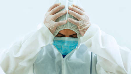 Front view of doctor with blue mask and bioprotective suit with hands on head tilted down with attitude of tiredness, sadness, crying or worry on white backgroundの写真素材